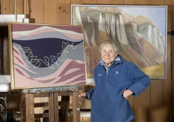 Doris McCarthy in a blue pullover, standing next to one of her abstract oil paintings on an easel. Another oil painting of cliffs hang on the wall behind her.